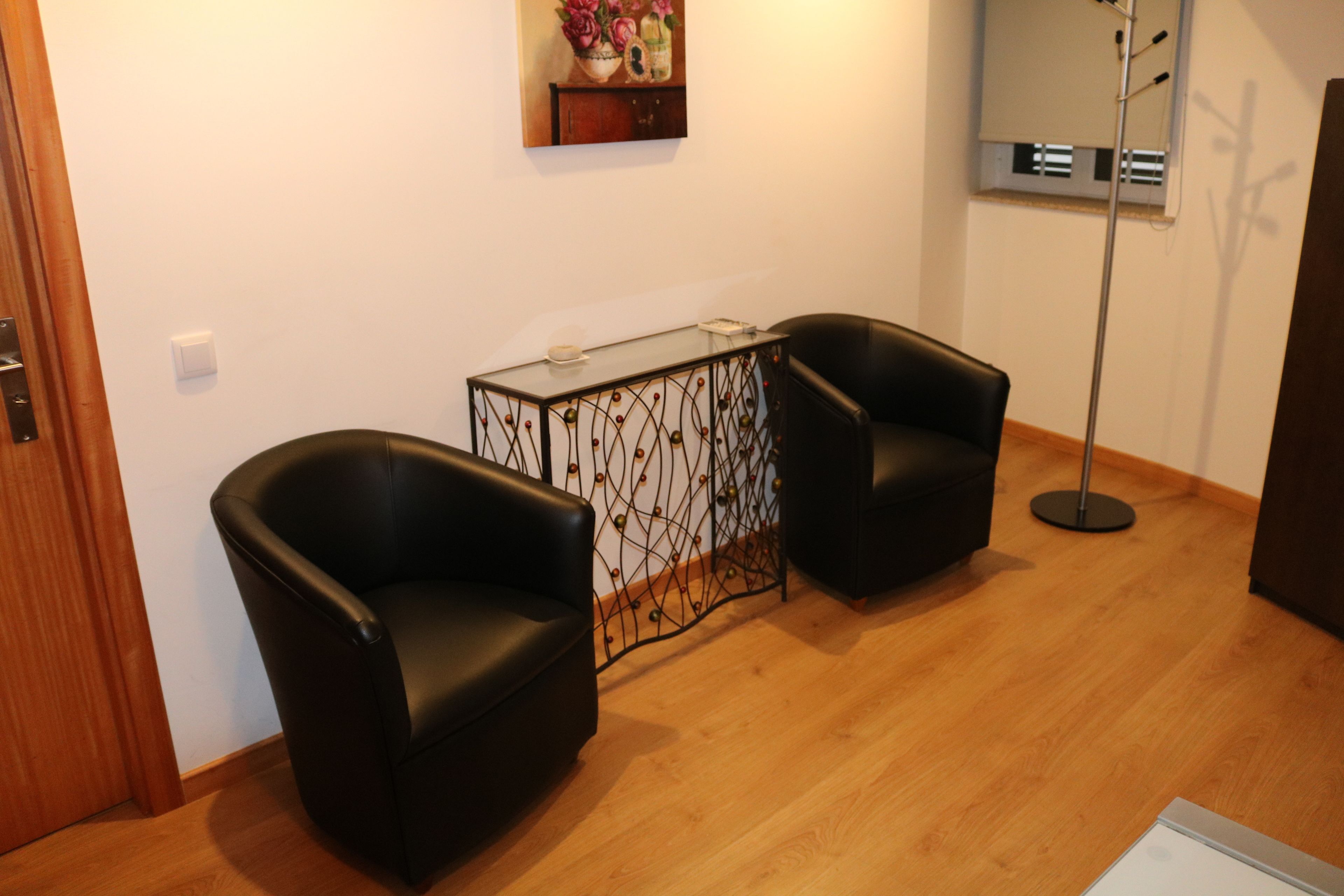 A room with two black leather chairs, a glass-top table with a decorative metal frame, a wooden floor, and a wall-mounted air conditioning unit.