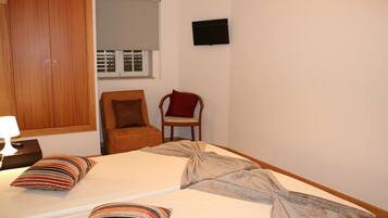 A bedroom with two beds, a wooden wardrobe, a chair, a TV, and a window with blinds.