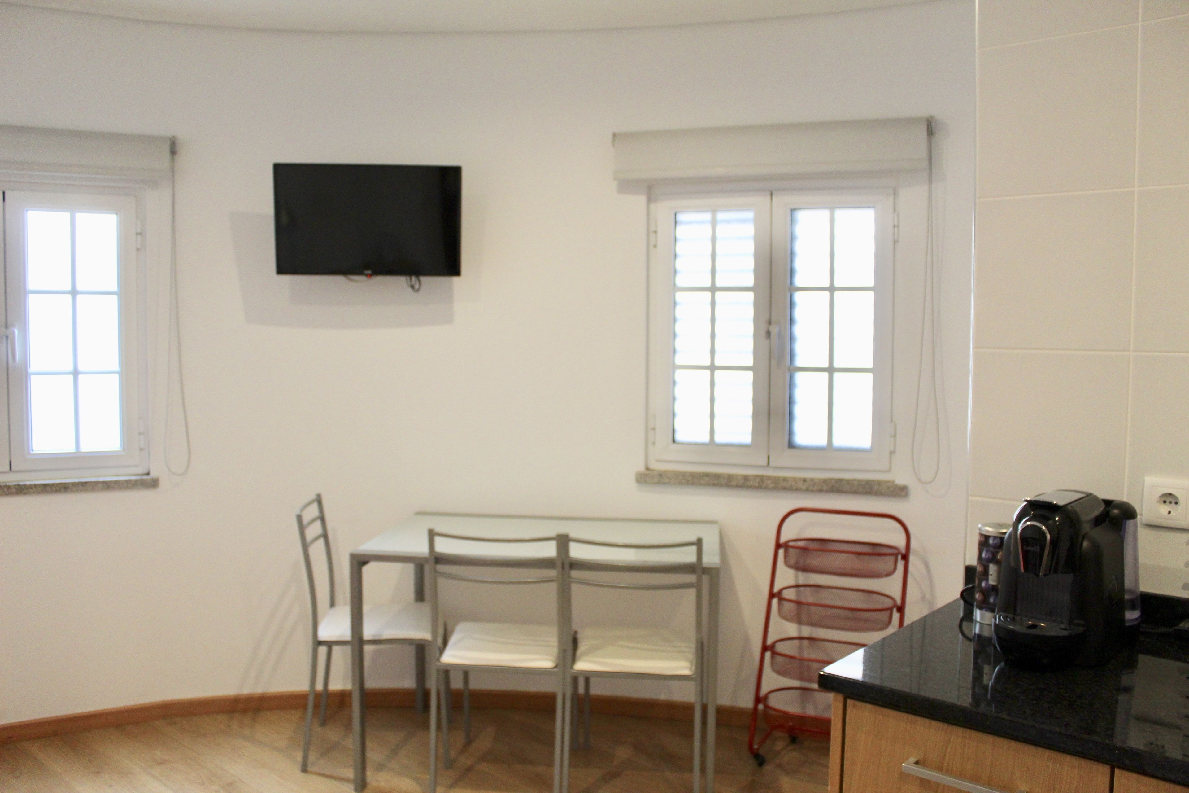 Una habitación pequeña y limpia con kitchenette, un televisor montado en la pared y dos ventanas que ofrecen luz natural.