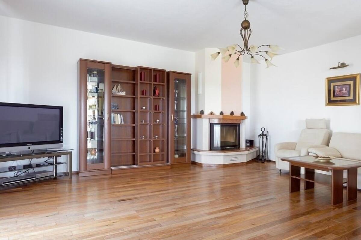 A living room with a flat-screen TV, wooden flooring, a sofa, a coffee table, a fireplace, and a bookshelf.