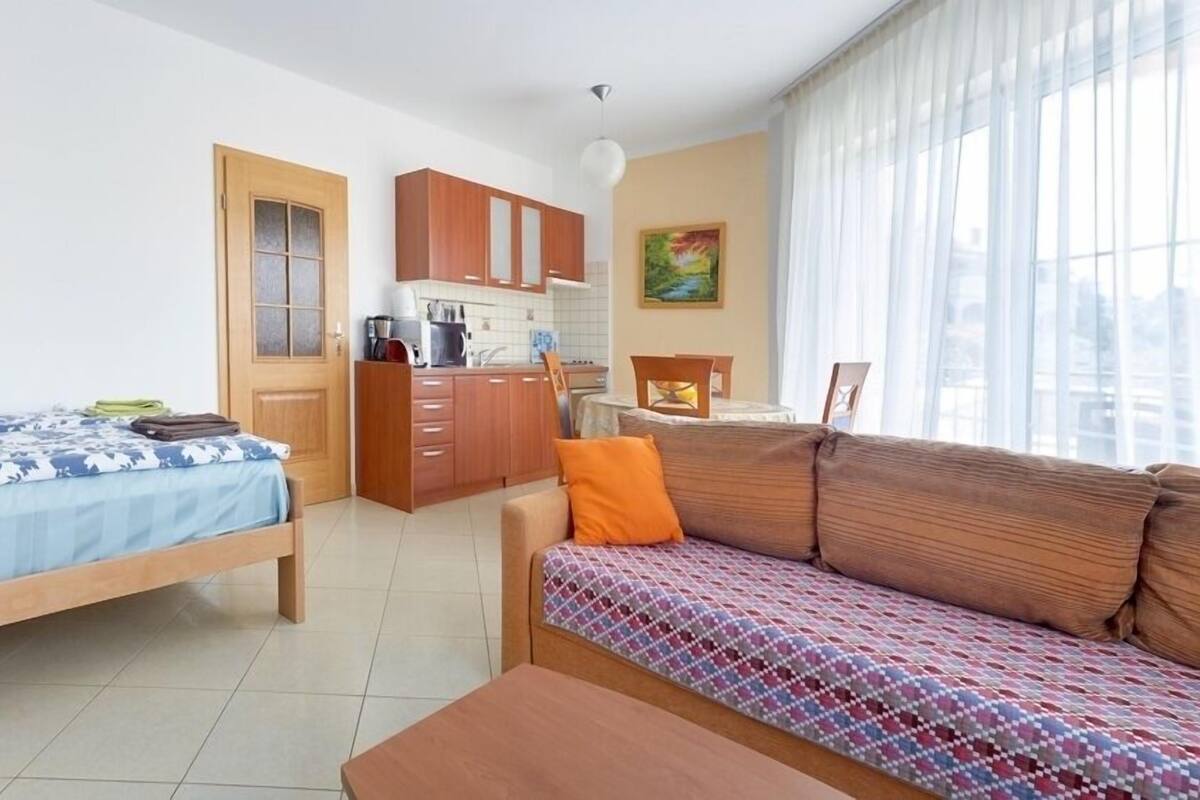 A compact room with a bed, a sofa, a small table, a kitchenette, and a window with curtains.