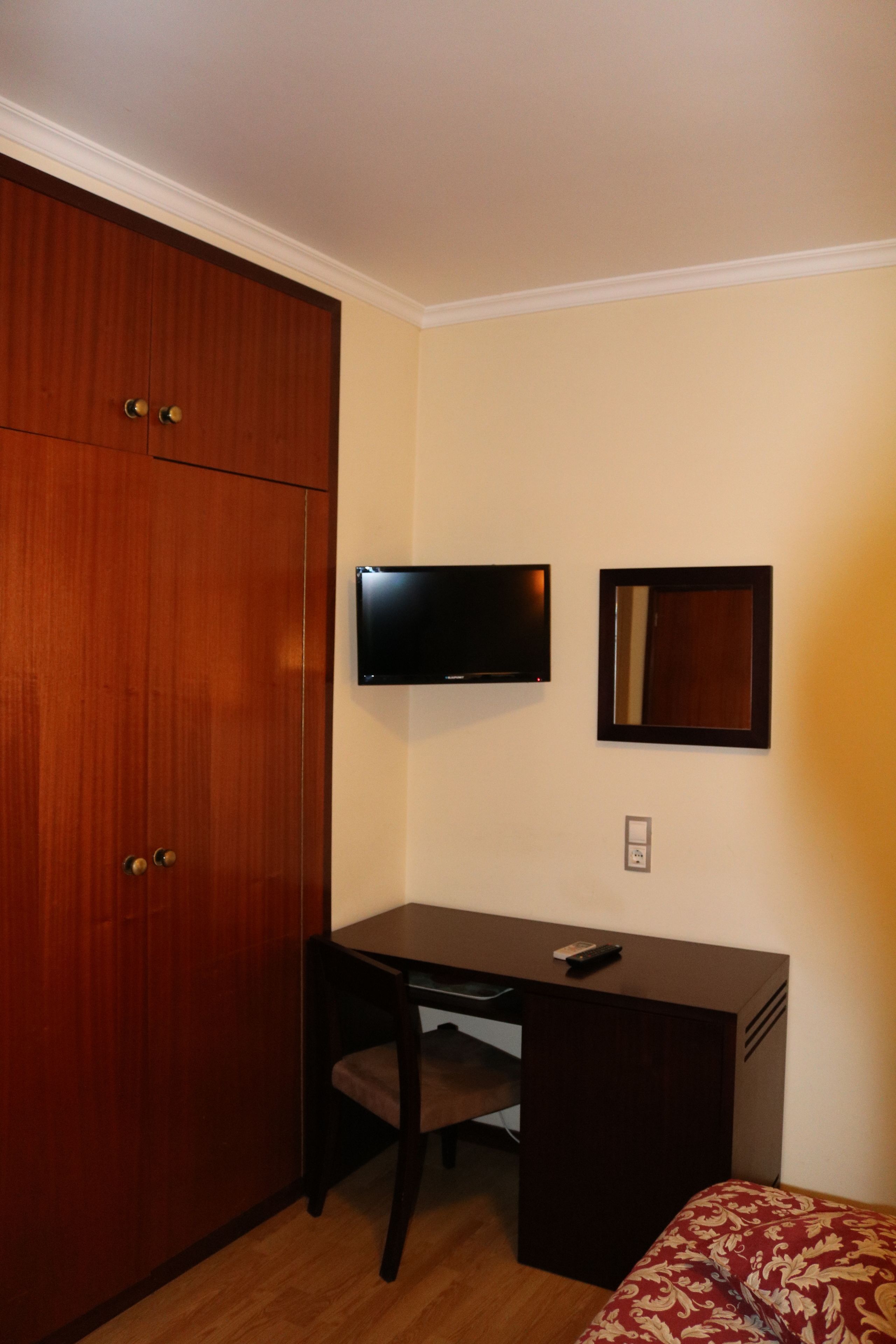 Una habitación con un armario de madera, un escritorio con silla, un televisor montado en la pared y un espejo pequeño.