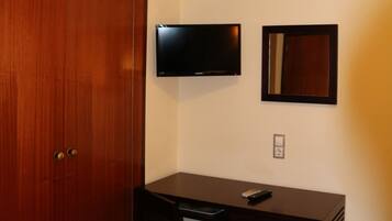 Una habitación con un armario de madera, un escritorio con silla, un televisor montado en la pared y un espejo pequeño.