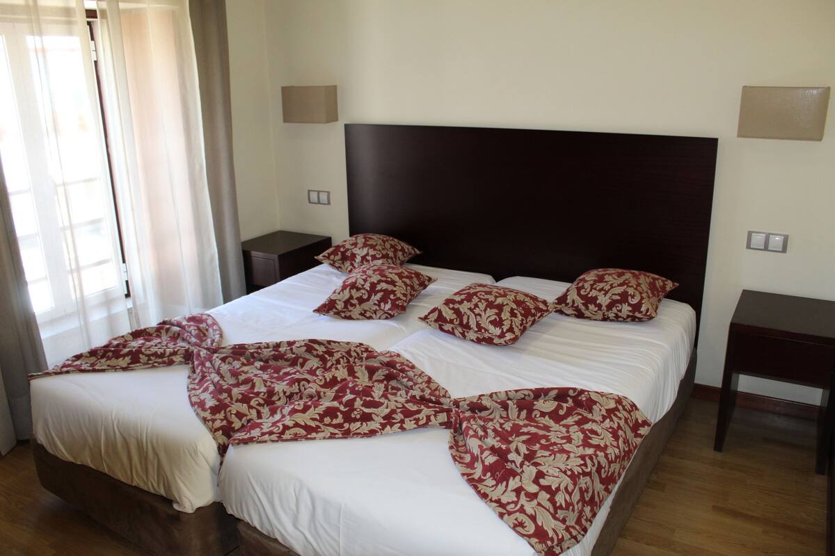 A neatly made bed with a white and red floral bedspread, two bedside tables, and a window with sheer curtains.