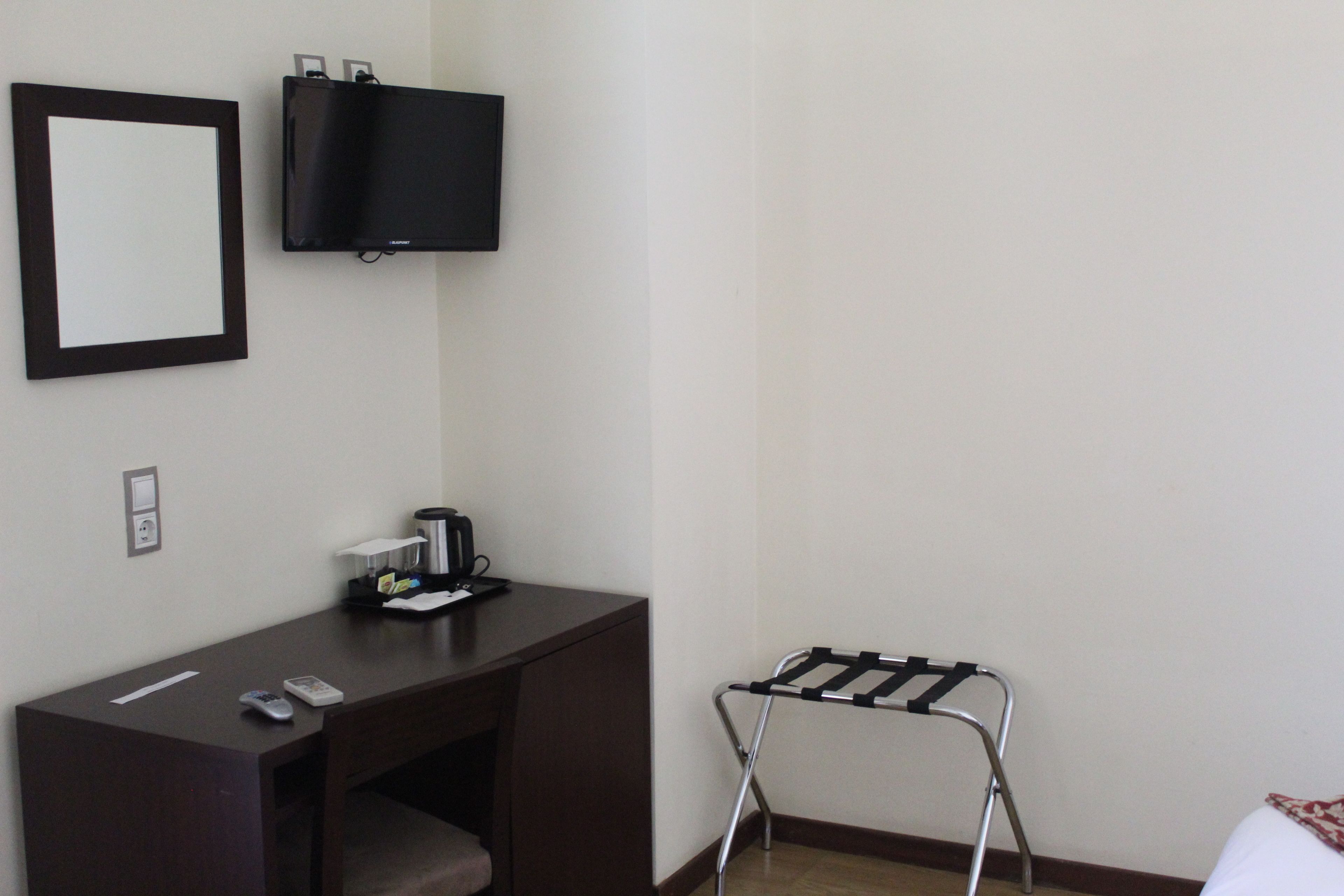 A hotel room with a desk, a TV mounted on the wall, a small table with a coffee maker, and a portable chair.