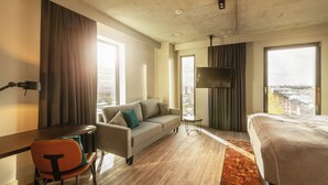 Habitación de hotel con sofá, cama, escritorio y silla. Hay un televisor sobre un mueble bajo y una ventana con vista a la ciudad.