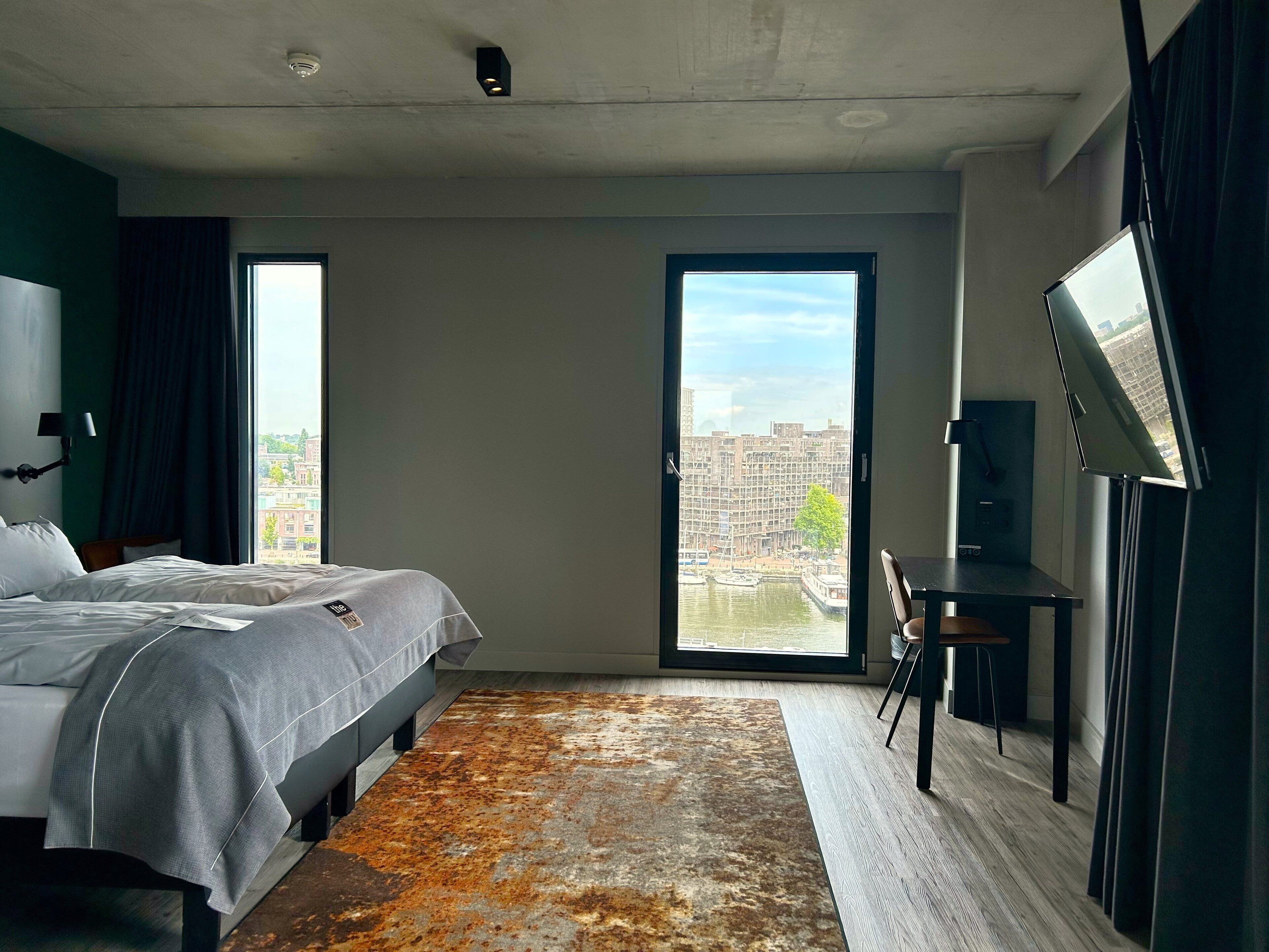 A modern hotel room with a large bed, a desk, and a view of the city through a large window.