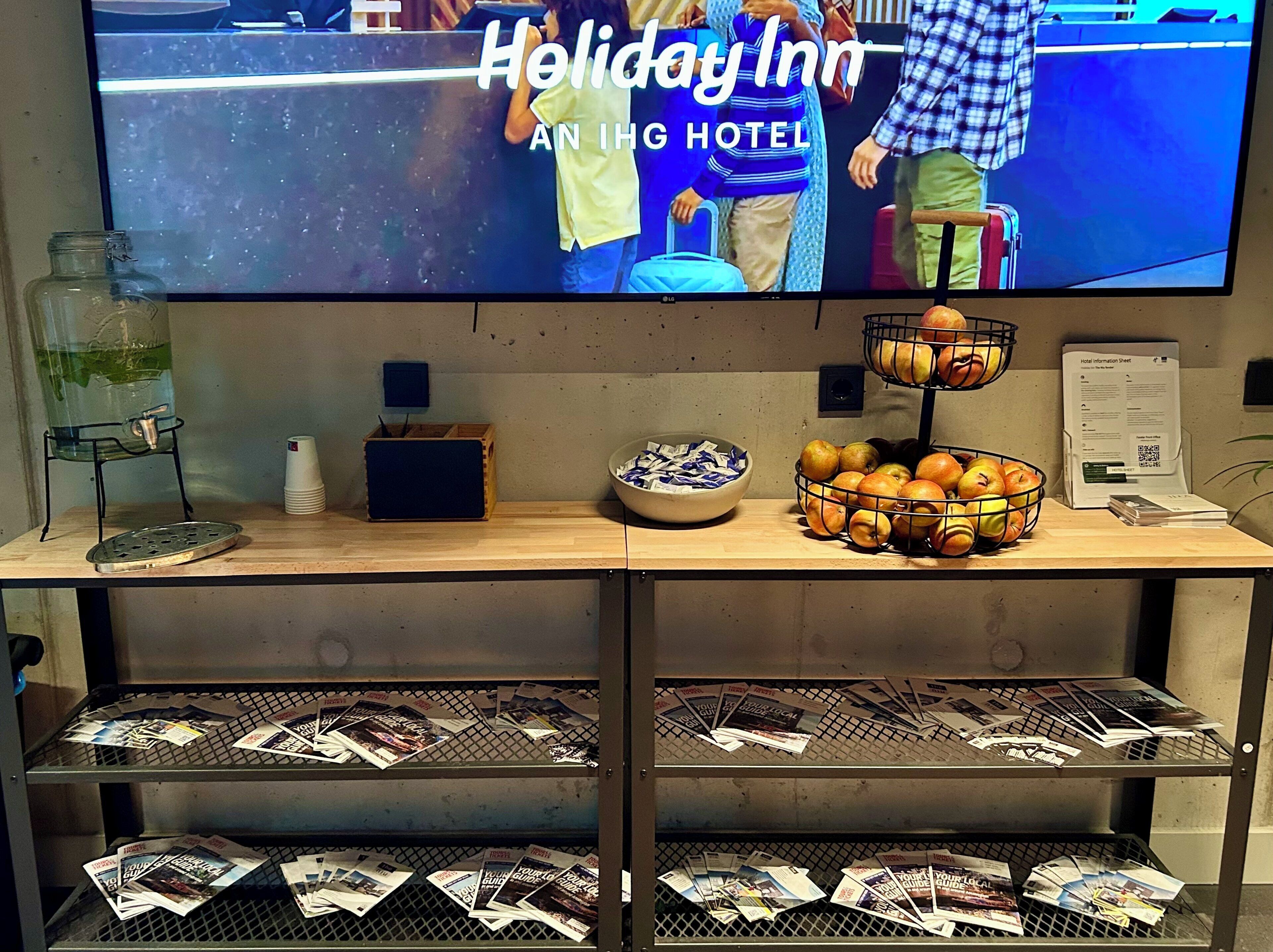 Un espace d’accueil d’hôtel avec un téléviseur affichant les logos « Holiday Inn » et « An IHG Hotel », un panier de fruits, un distributeur d’eau, une pile de magazines et une fiche d’information sur l’hôtel.