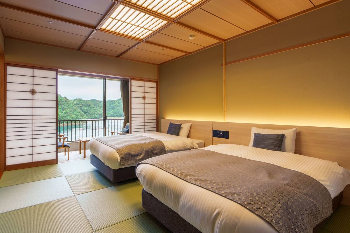 A hotel room with two beds, tatami flooring, a sliding door to a balcony, and a view of a mountainous landscape.