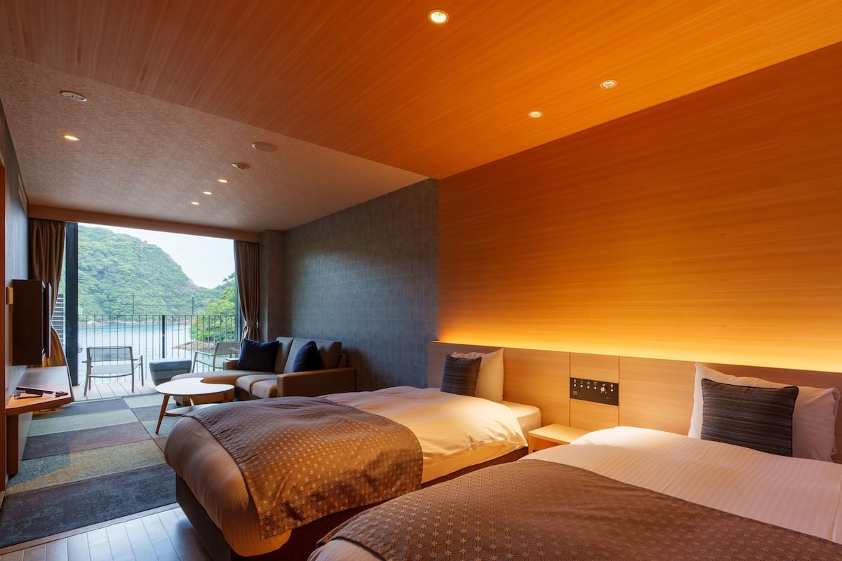 A hotel room with two beds, a sofa, a small table, and a TV. There is a balcony with a view of the sea.
