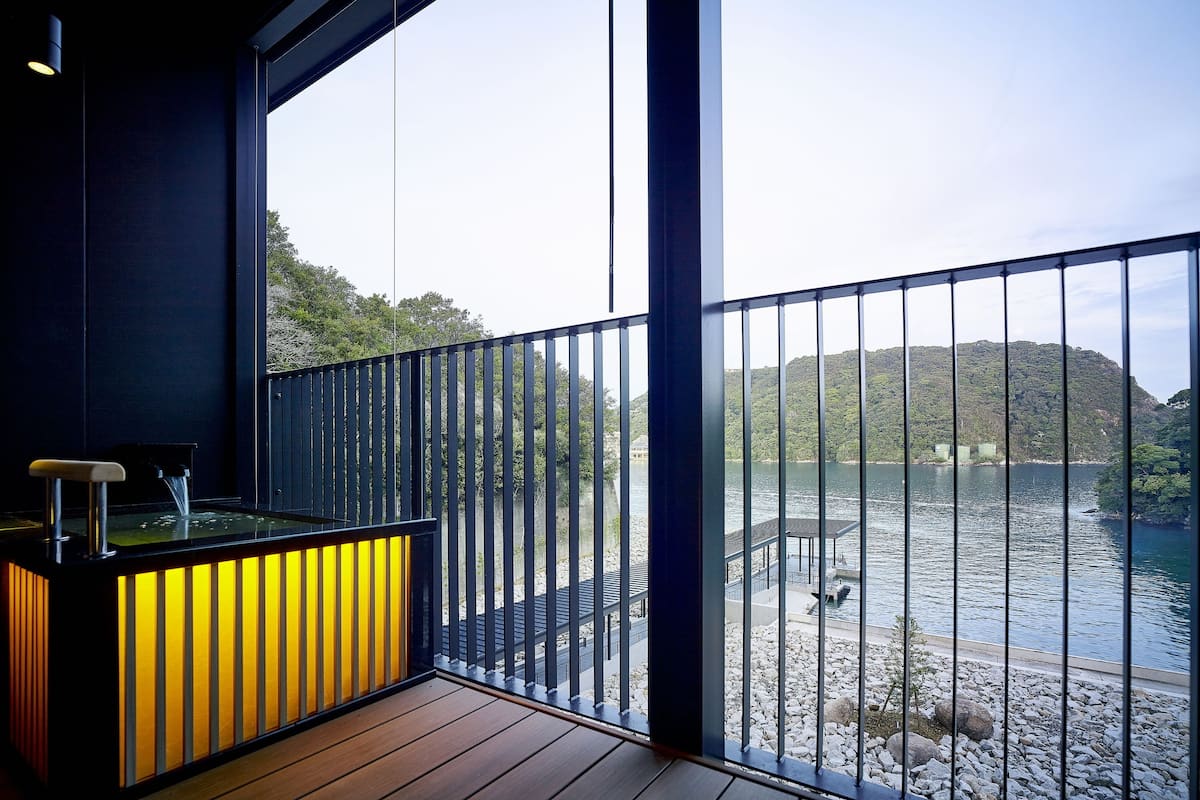 A balcony with a view of a lake and hills, a wooden floor, and a bar area with a yellow-lit cabinet.