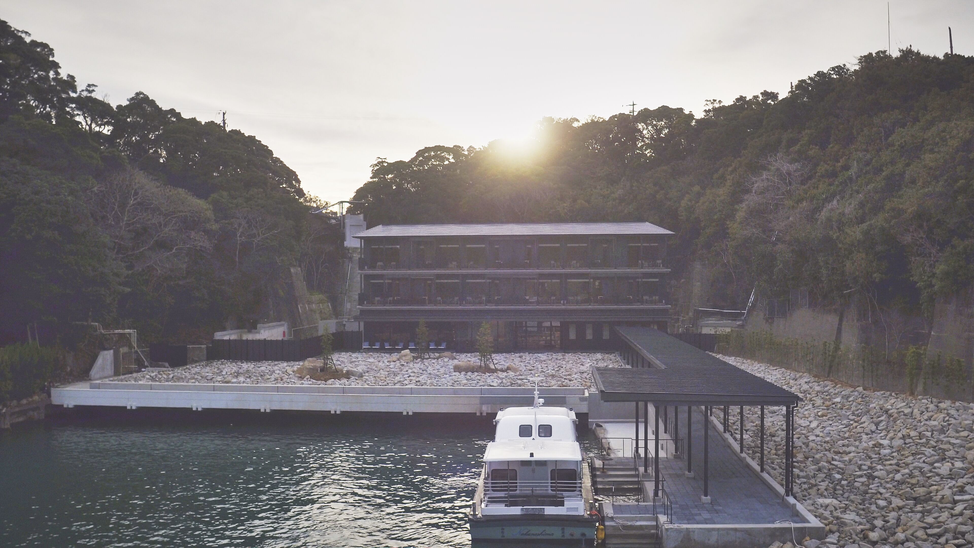 Un edificio moderno de varios pisos con un muelle que se adentra en una masa de agua tranquila, rodeado de terreno rocoso y vegetación densa.