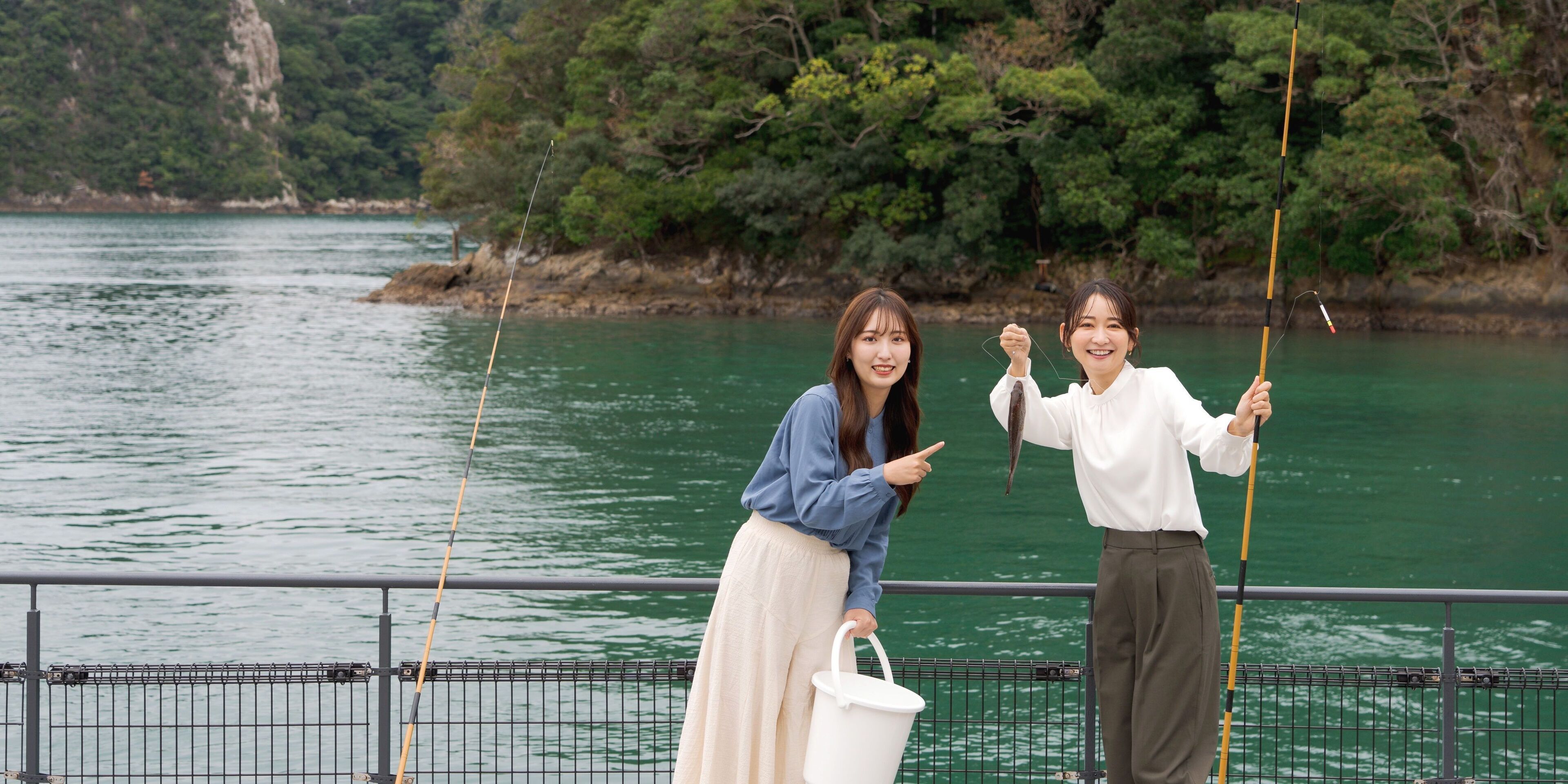 Two people fishing by a railing with a body of water and trees in the background.