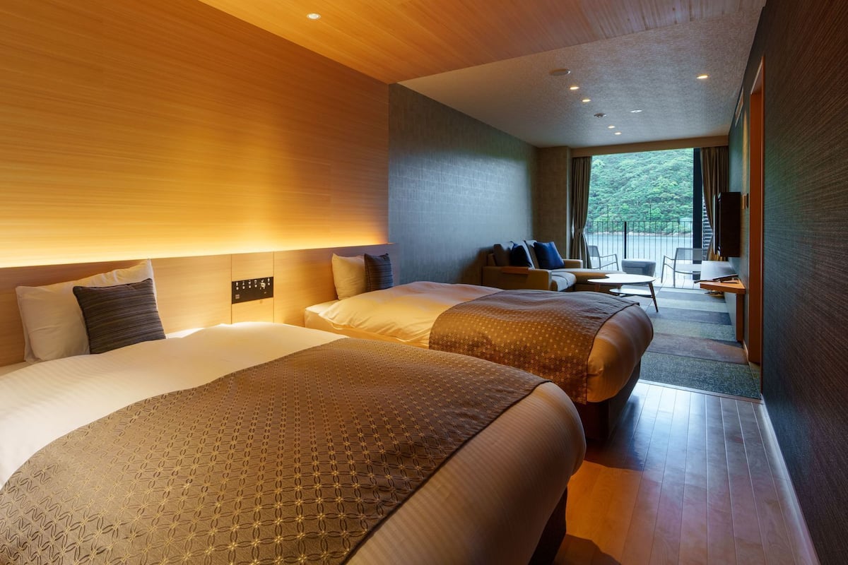 A modern hotel room with two beds, a sofa, a desk, and a balcony with a view.