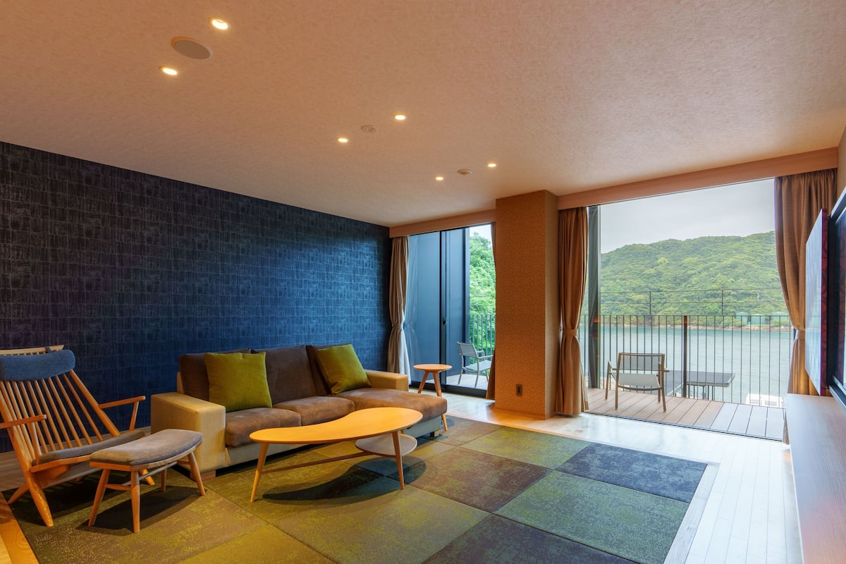 A modern living room with a blue textured wall, a sofa with green cushions, a wooden chair, a small round table, and a balcony with a view of greenery.