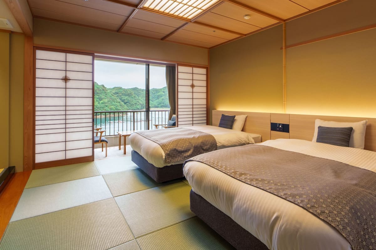 A hotel room with two beds, tatami flooring, a balcony with a view, and a traditional Japanese sliding door.