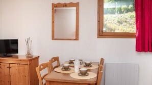 Un coin repas avec une table en bois, quatre chaises, un miroir, une fenêtre avec un rideau rouge, un radiateur et un téléviseur posé sur une commode en bois.