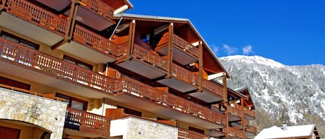 A multi-story building with wooden balconies and stone walls, set against a snowy mountain backdrop.
