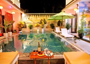 A poolside dining area with a table set for two, a bottle of wine, and a plate of fruit. - Green Island Hoian Villa (Da Nang)