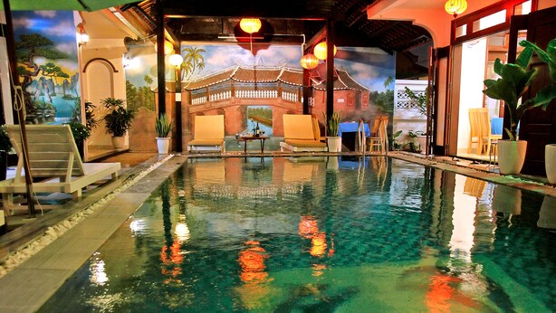 Una piscina interior con agua cristalina, un mural de una edificación tradicional y varias sillas de descanso.