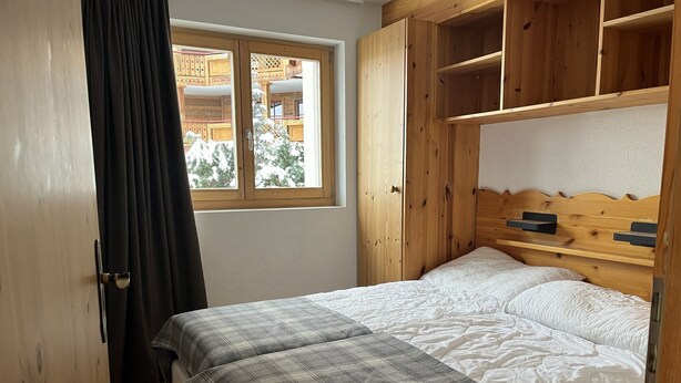 A bedroom with a wooden headboard, a bed with white bedding, a plaid blanket, and a window with a view of a wooden building outside. - Pracondu 1 002 - OUTDOOR & FUN  charming apartment (Haute-Nendaz)
