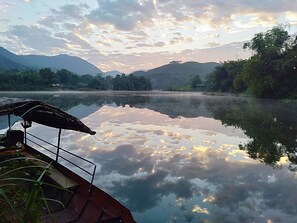 A calm lake with a boat docked on the left, surrounded by lush greenery and mountains in the background. - Vu Linh Farmstay - Hostel (Thac Ba)