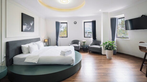 A modern hotel room with a curved bed, a flat-screen TV, a small round table, and a potted plant. - N Queen Apartment Hotel (Xuan Huong - Da Lat)