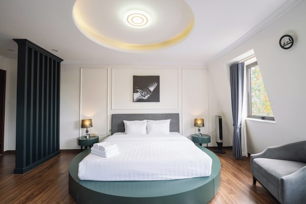 A modern hotel room with a round bed, two bedside tables, a standing fan, a grey armchair, and a window with curtains.