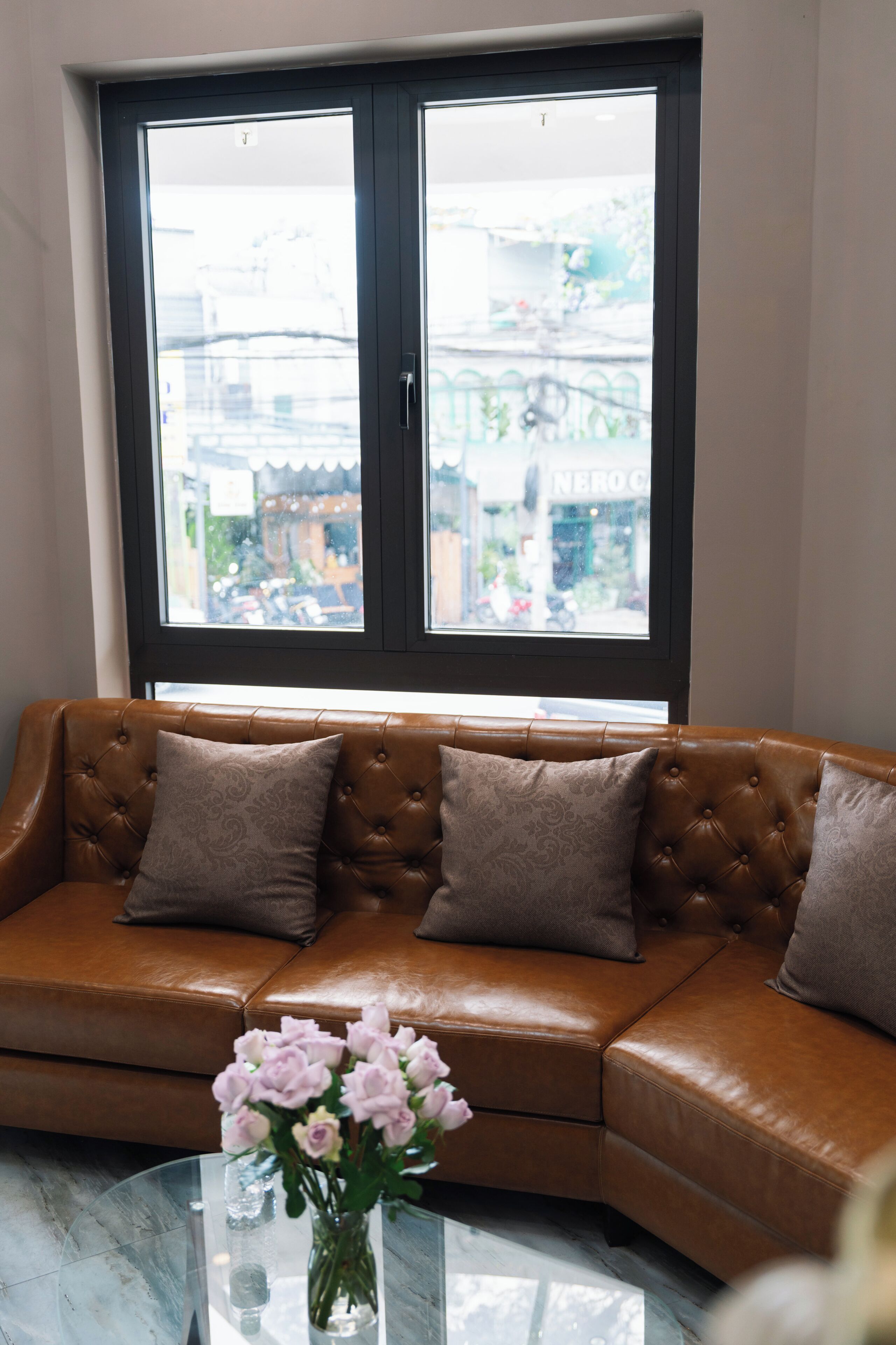 A brown leather sofa with patterned cushions, a glass coffee table with a vase of pink flowers, and a large window with a view of buildings and a street.