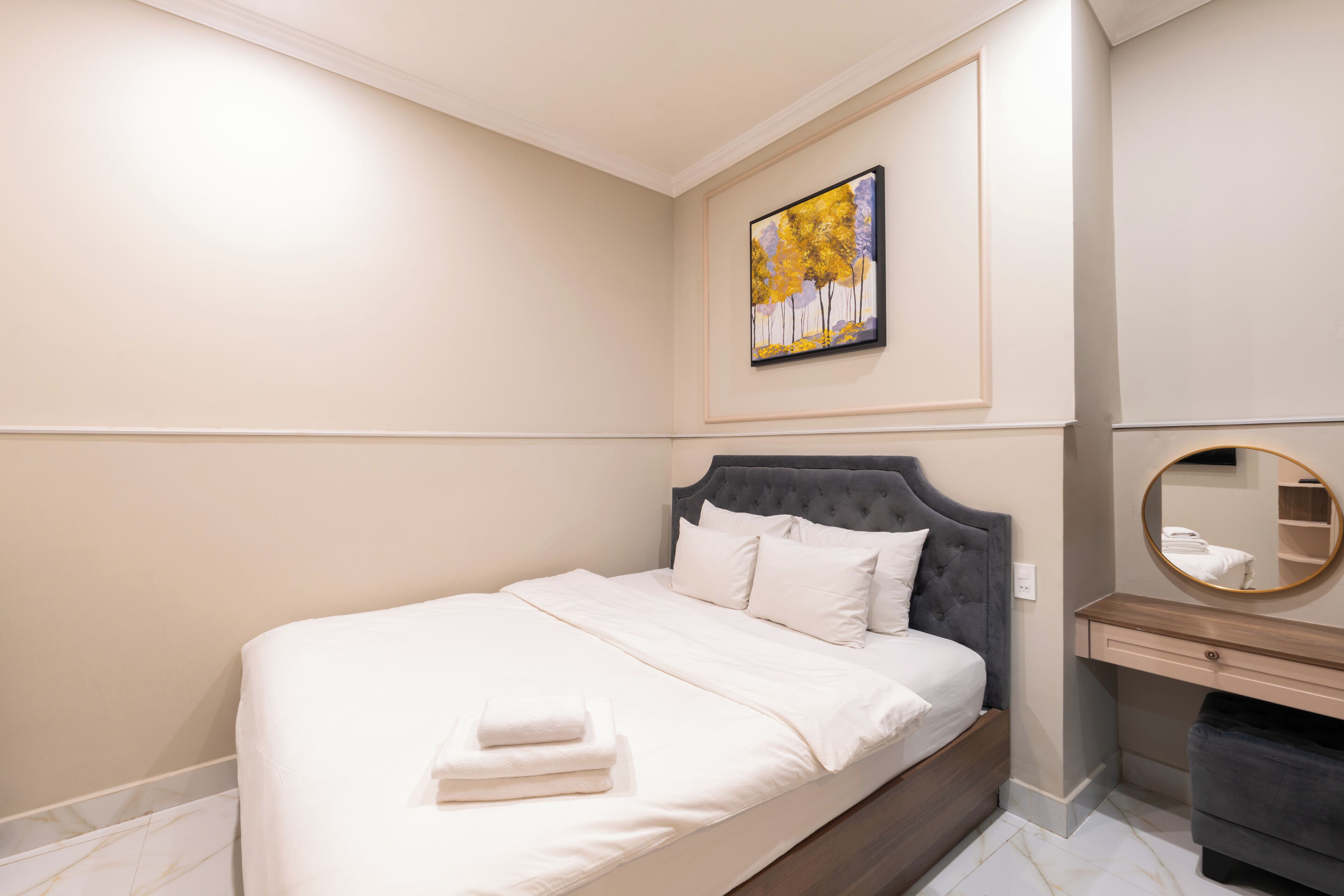 A neatly made bed with white linens and a folded towel, a round mirror above a wooden dresser, and a framed painting on the wall.