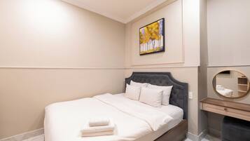 A neatly made bed with white linens and a folded towel, a round mirror above a wooden dresser, and a framed painting on the wall.