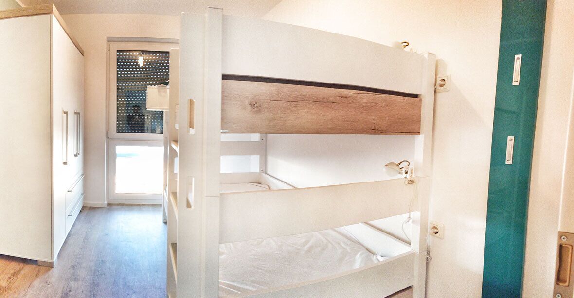 Een stapelbed met een houten hoofdbord, een witte matras en een metalen ring aan de zijkant. Er is een deur met een groene hendel en een witte muur met een raam.