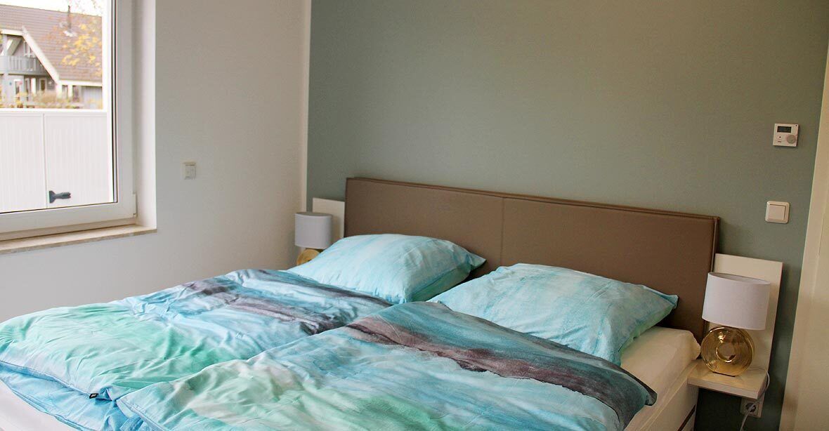 Un dormitorio con cama, dos lámparas de noche y ventana con vista a una casa.