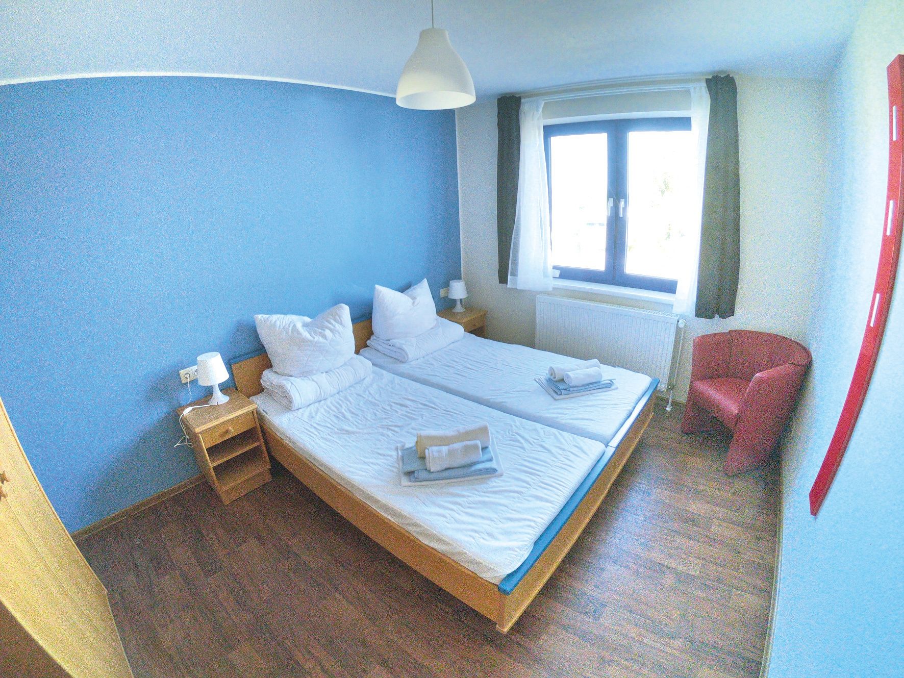 Un lit double avec du linge de lit blanc, une table de chevet avec une lampe, une fenêtre avec des rideaux et un fauteuil rouge.