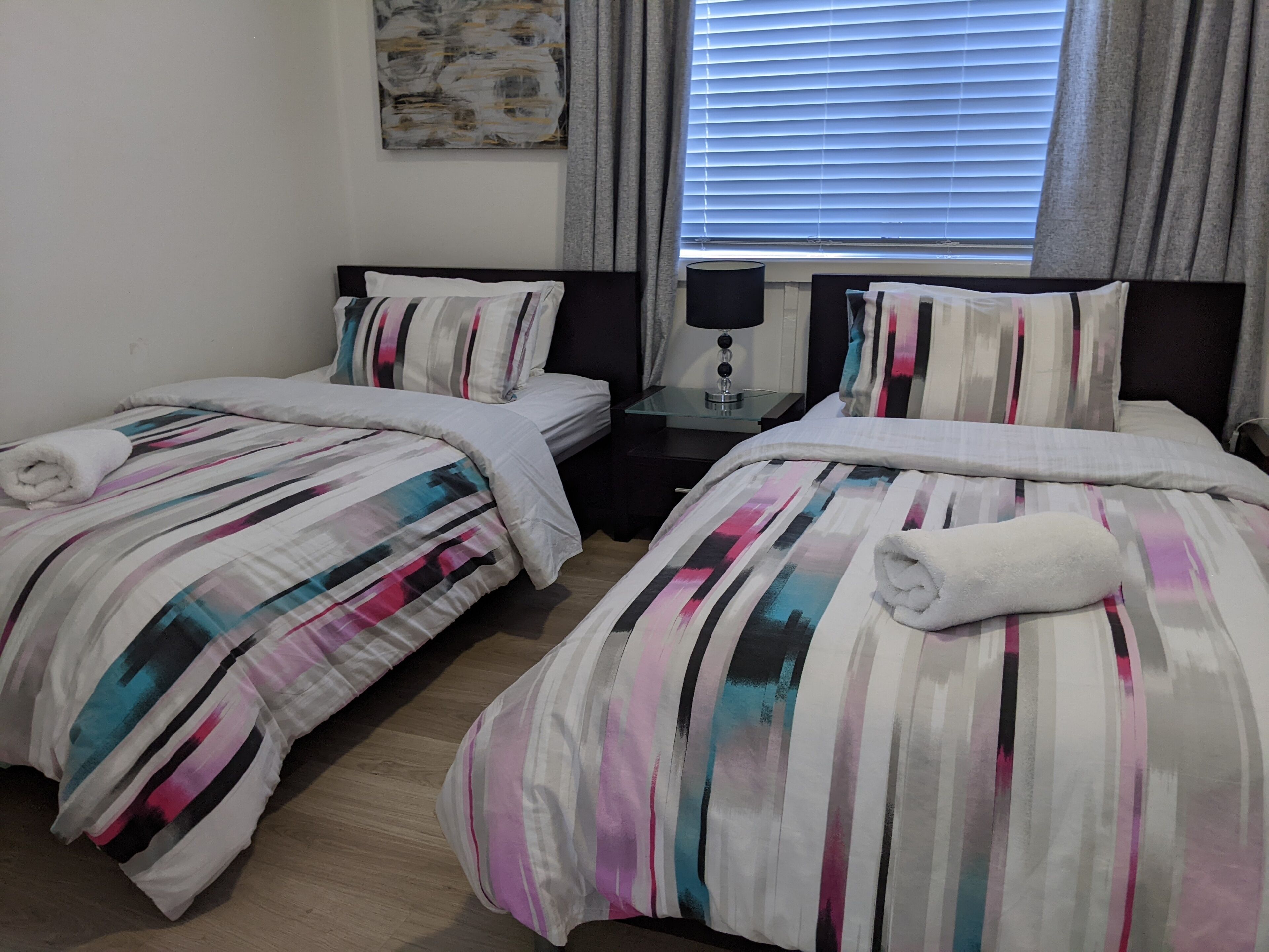 Ein Hotelzimmer mit zwei Betten, jeweils mit gestreifter Bettwäsche und aufgerollten Handtüchern. Dazwischen befindet sich ein Nachttisch mit einer Lampe, und es gibt ein Fenster mit Jalousien.