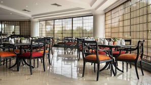 Uma sala de jantar ampla com várias mesas e cadeiras, grandes janelas com persianas e piso polido.