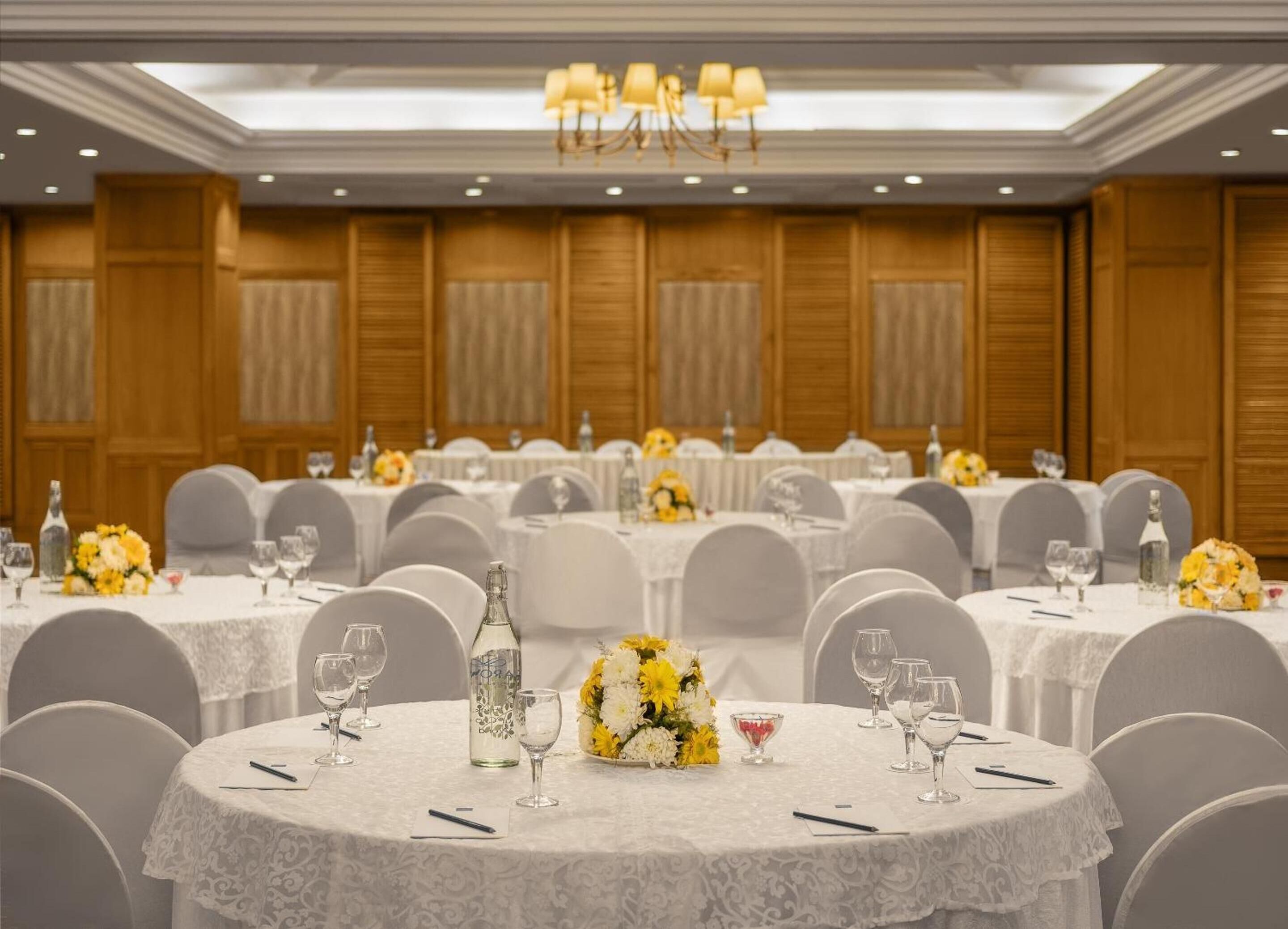 A banquet hall with round tables covered in white tablecloths, arranged in rows. Each table has a centerpiece of yellow and white flowers, wine glasses, and water bottles. The room has wooden paneling on the walls and a chandelier hanging from the ceiling.