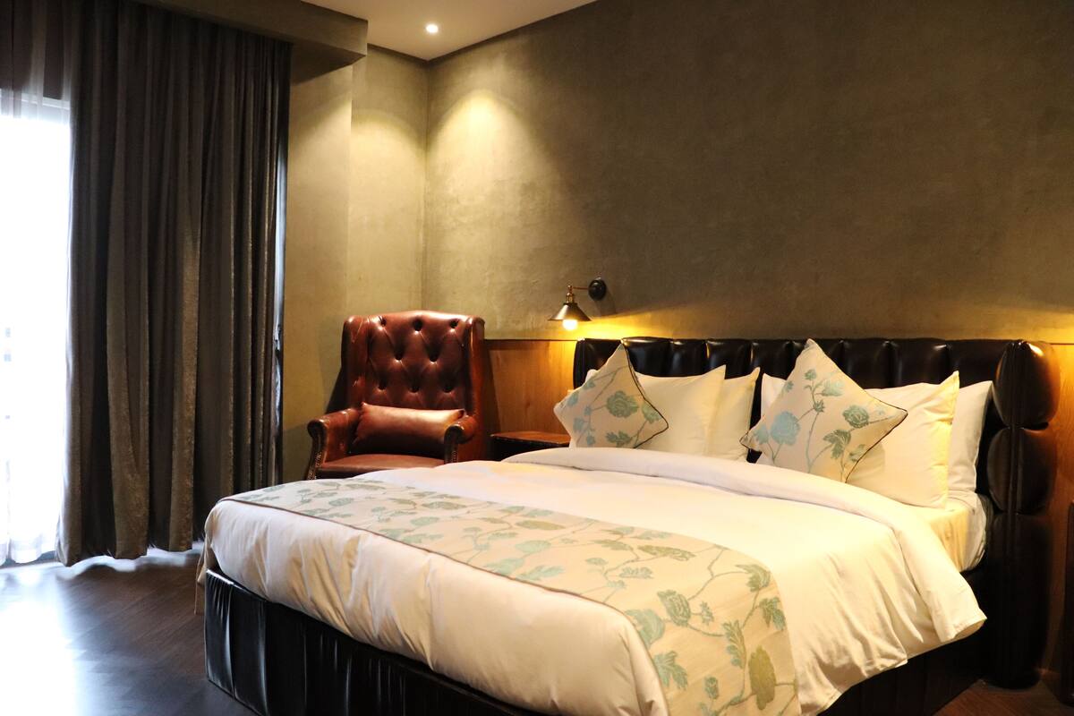 A hotel room with a large bed, a leather armchair, and a nightstand with a lamp.