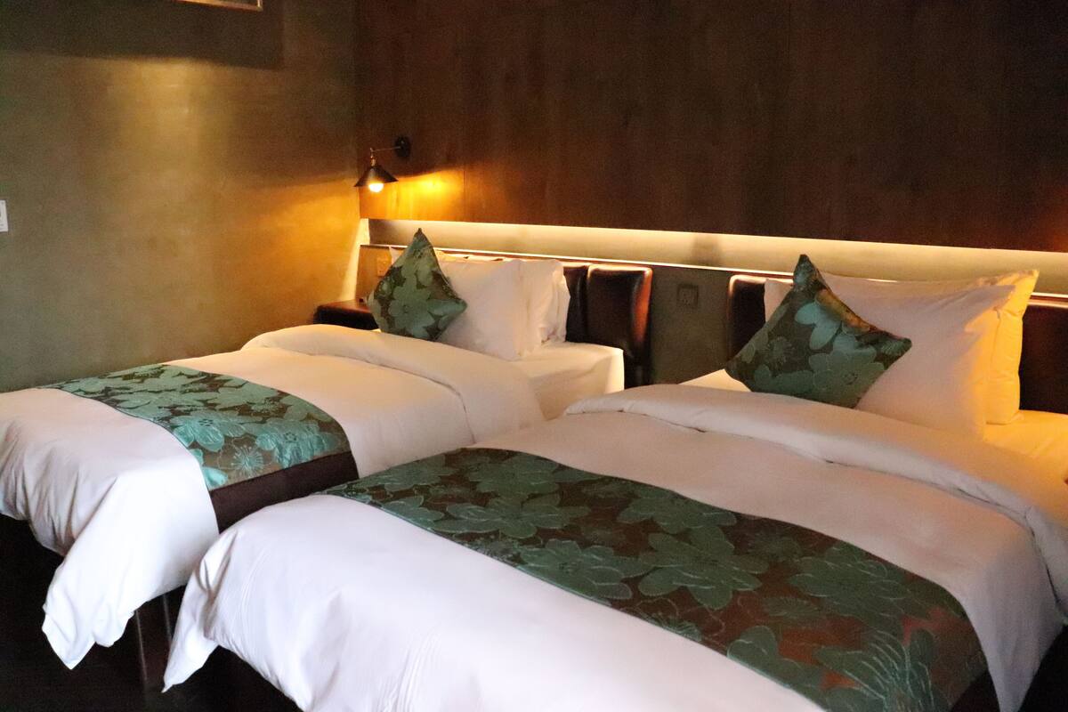 A hotel room with two beds, each with a green leaf-patterned bedspread and white pillows. There is a nightstand with a lamp between the beds.