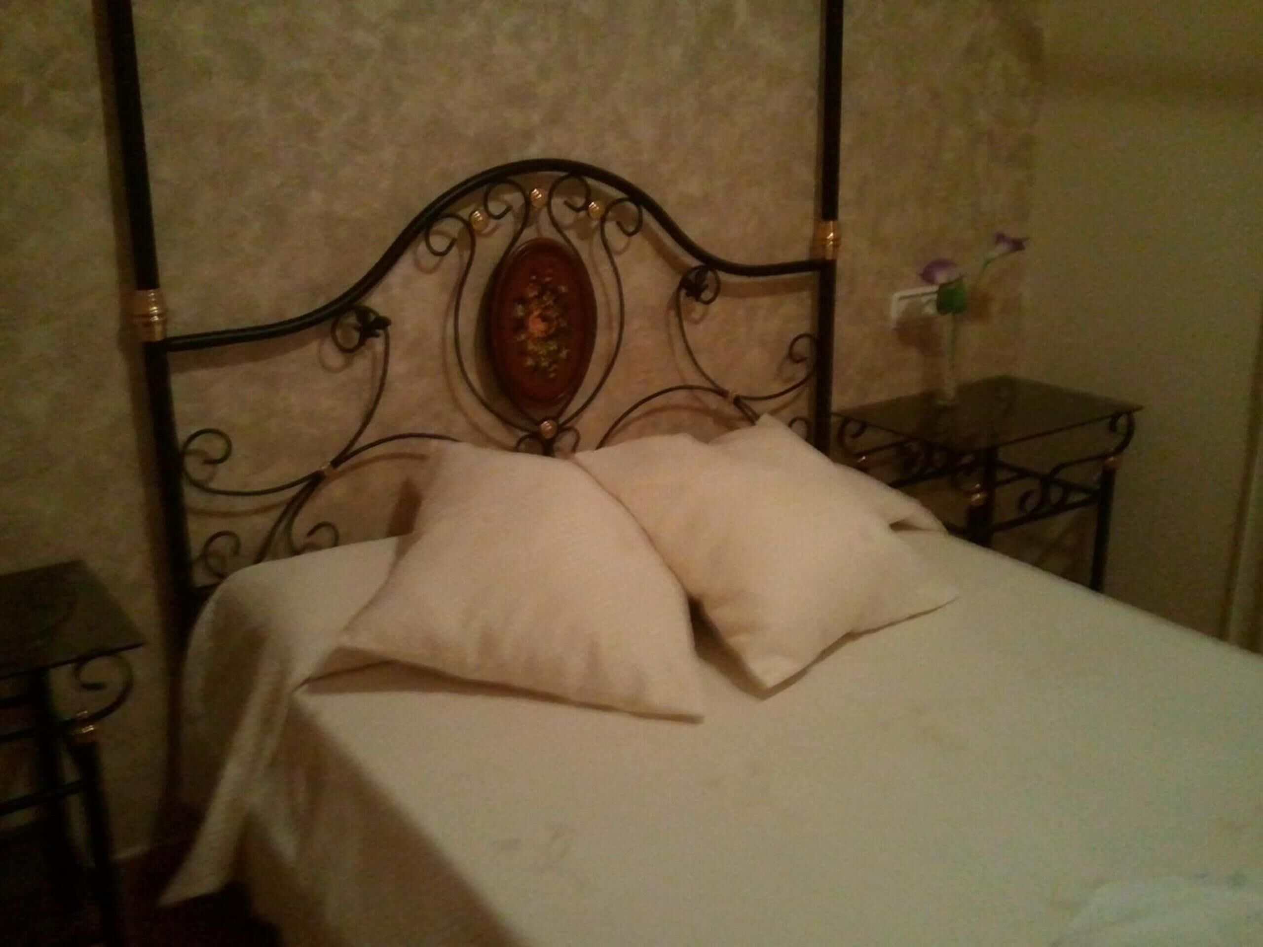 A bed with a metal headboard and two pillows, a small table with a vase of flowers, and a plain wall.