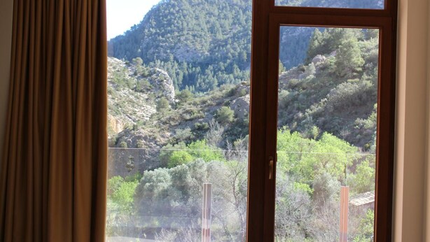 A window with a view of a mountainous landscape, curtains partially drawn, and a wooden frame. - Ca L'angels (Prat de Comte)