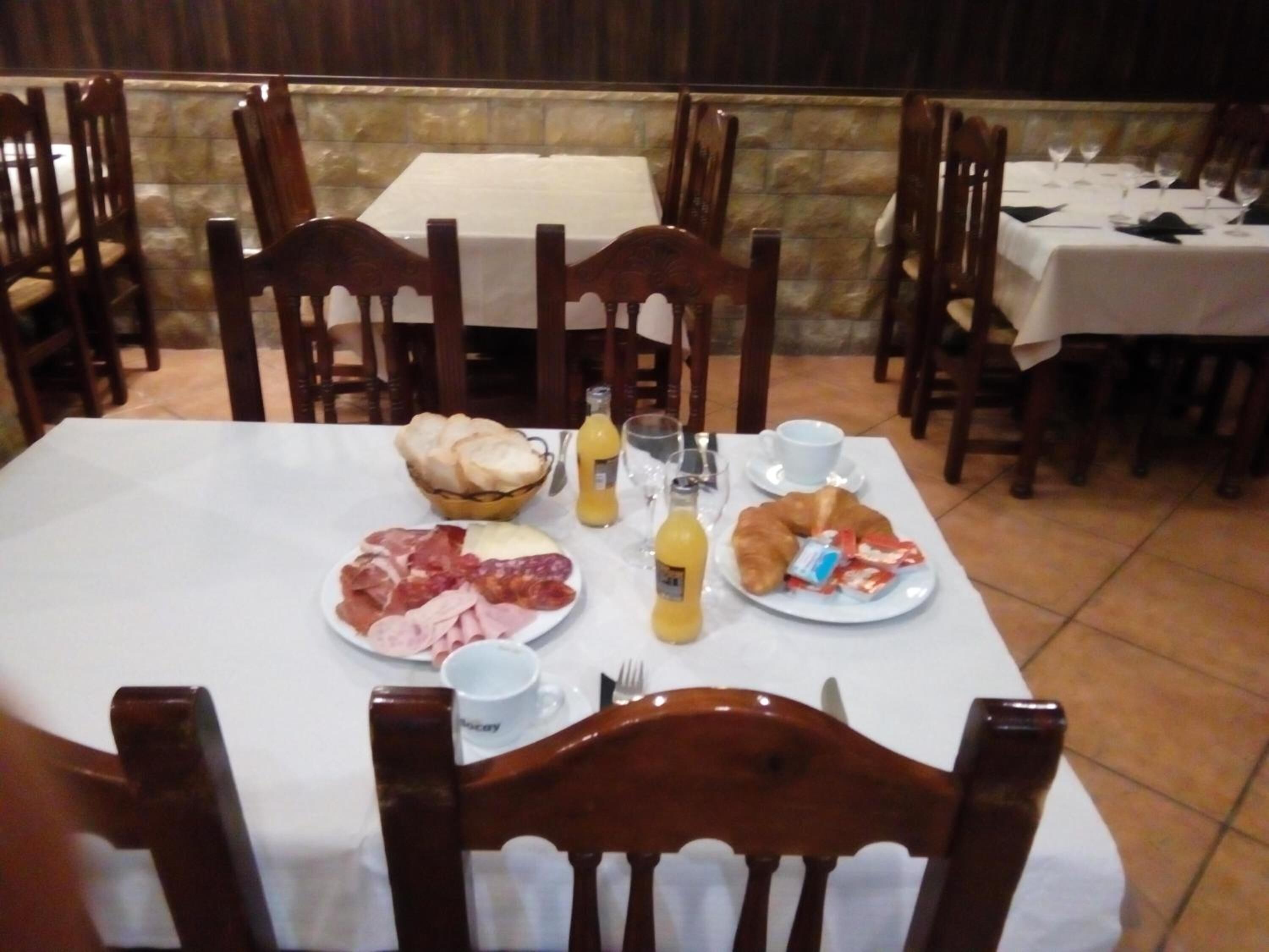 A dining table set with breakfast items, including bread, ham, and croissants, in a restaurant with wooden chairs and a stone wall background.