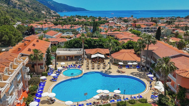 A resort with a large swimming pool surrounded by lounge chairs and umbrellas, set against a backdrop of coastal town buildings and a clear blue sea. - Ölüdeniz Turquoise Hotel (Fethiye)