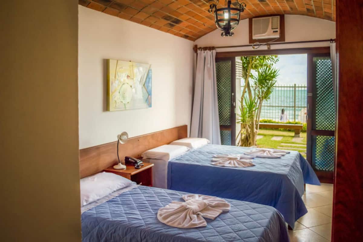 A hotel room with two beds, a wooden headboard, a nightstand, a lamp, and a view of the garden and sea through a glass door.