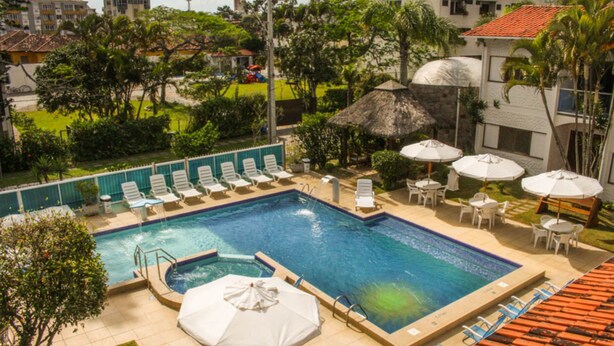 A swimming pool with lounge chairs and umbrellas, surrounded by greenery and buildings. - Village Paraiso Hotel (Florianópolis)