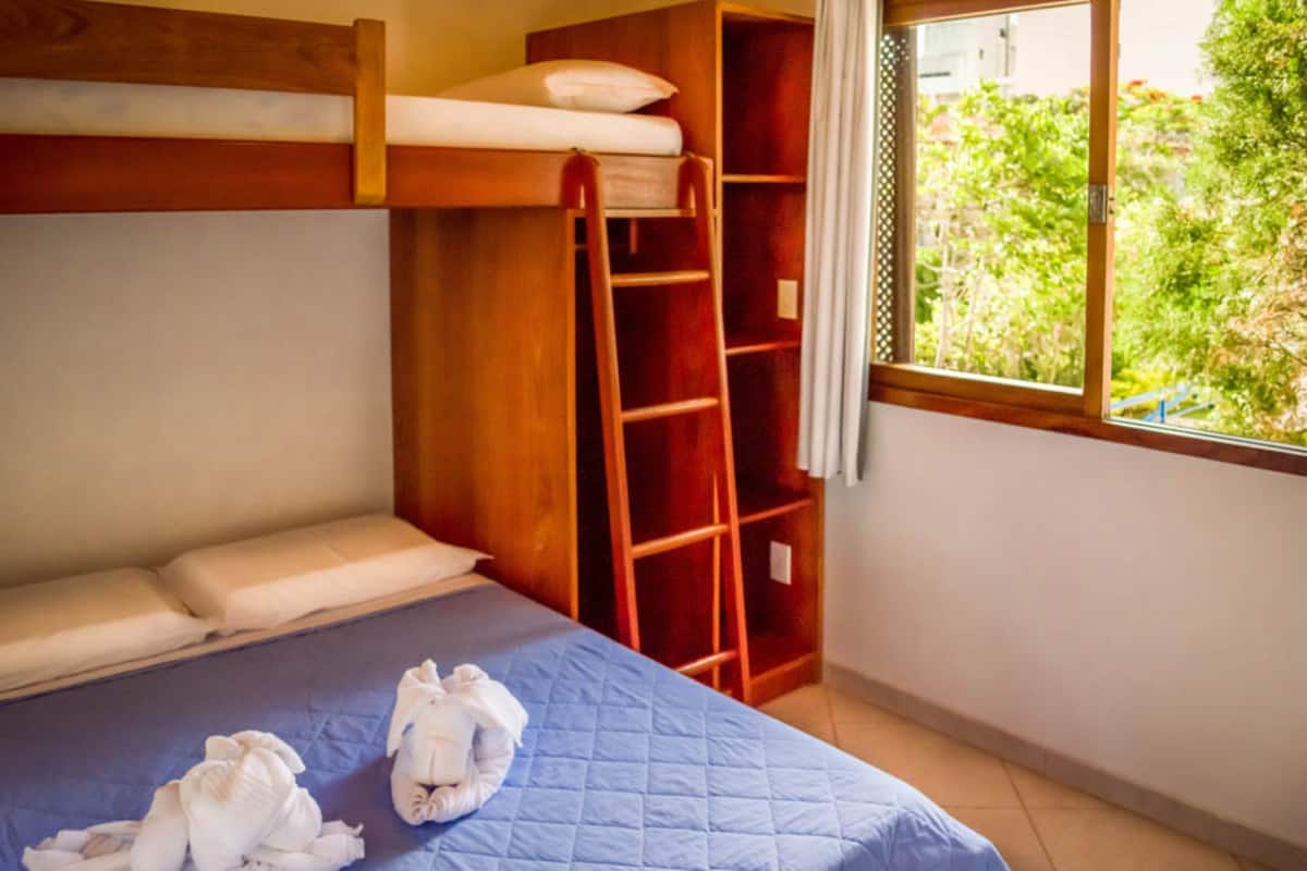 A bunk bed room with a ladder, a window with a view of greenery, and a neatly made bed with towels.