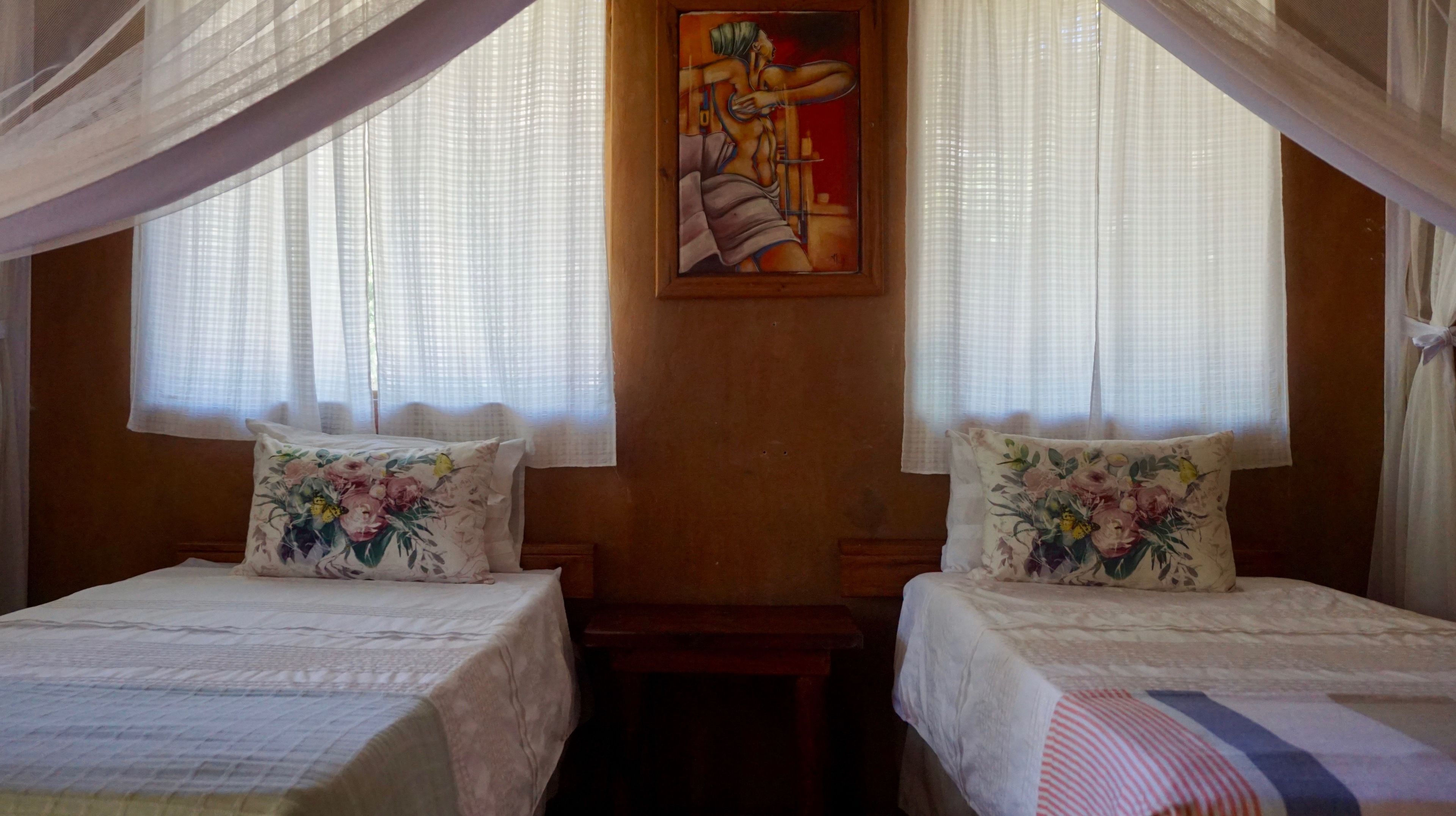 Una habitación con dos camas, cada una con almohadas de estampado floral, un cuadro en la pared y cortinas de encaje.