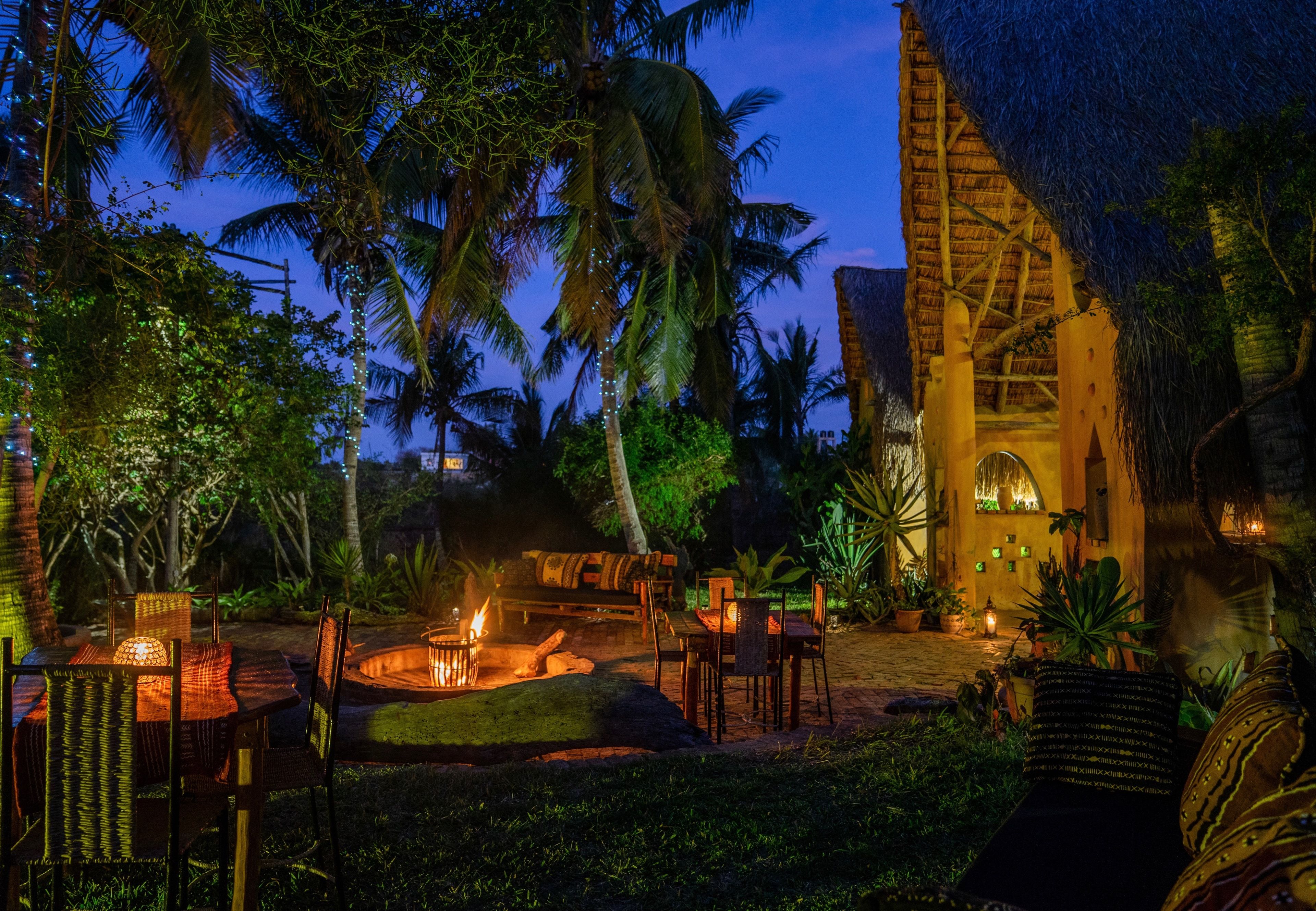 An outdoor dining area with a fire pit, tables, and chairs, surrounded by palm trees and a thatched-roof building.
