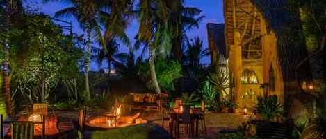 An outdoor dining area with a fire pit, tables, and chairs, surrounded by palm trees and a thatched-roof building.