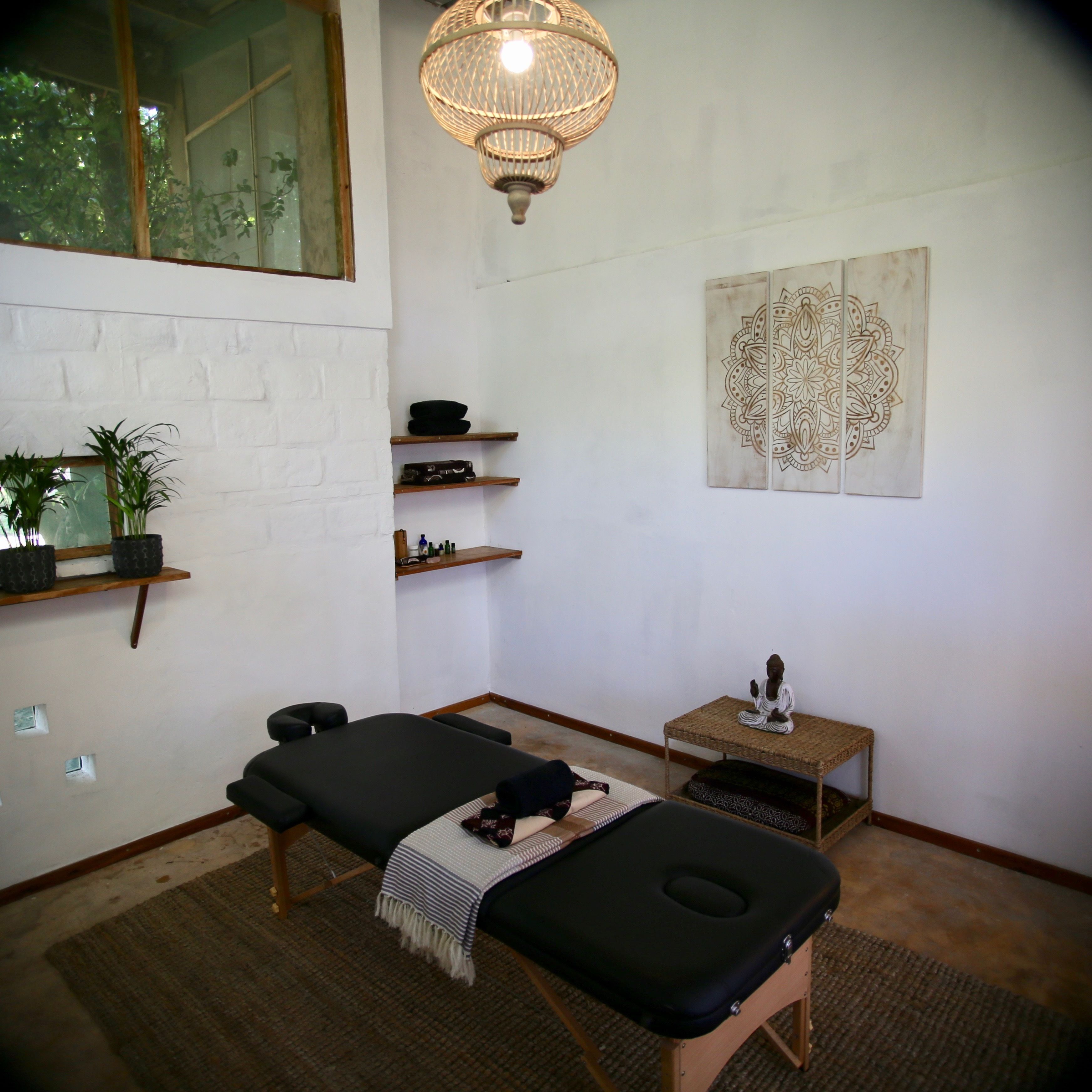 Quarto com mesa de massagem, prateleira pequena com objetos, peça decorativa na parede e luminária pendente.